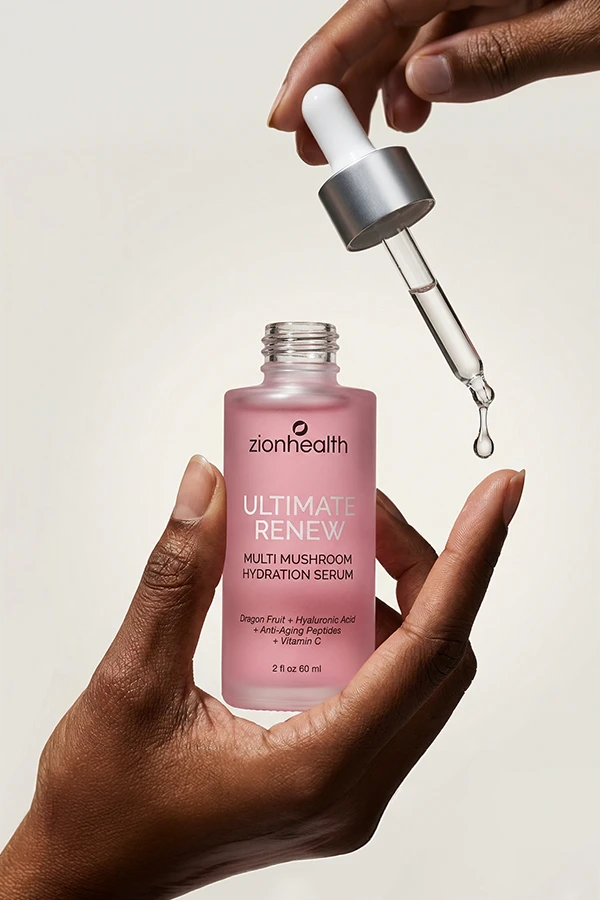 A dark-skinned hand holding a frosted pink bottle of Zionhealth Ultimate Renew Multi Mushroom Hydration Serum with an eye dropper.