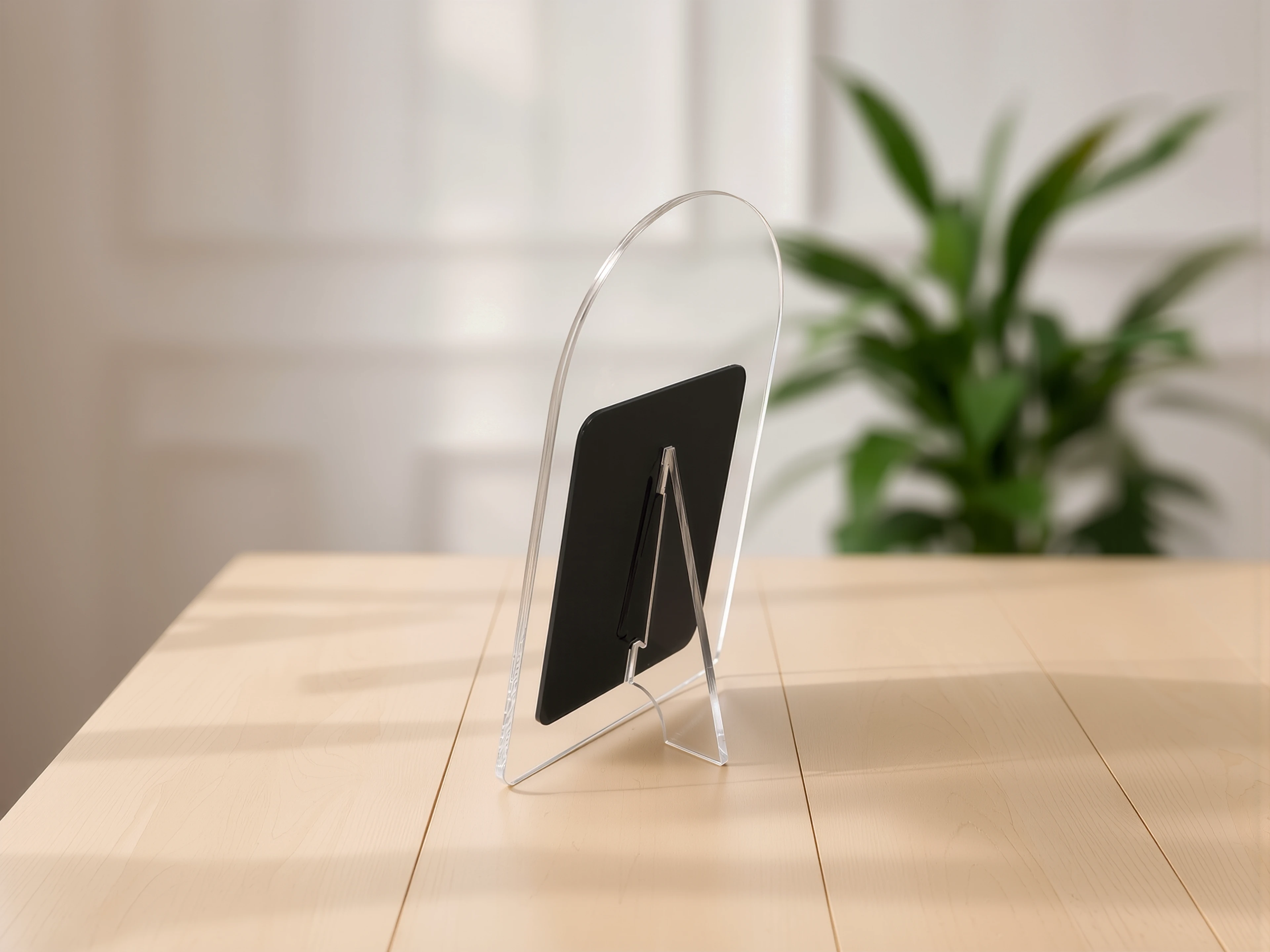 A clear acrylic MUUJEE sign stand holding a dark tablet-style insert, on a wood table beside a potted plant.