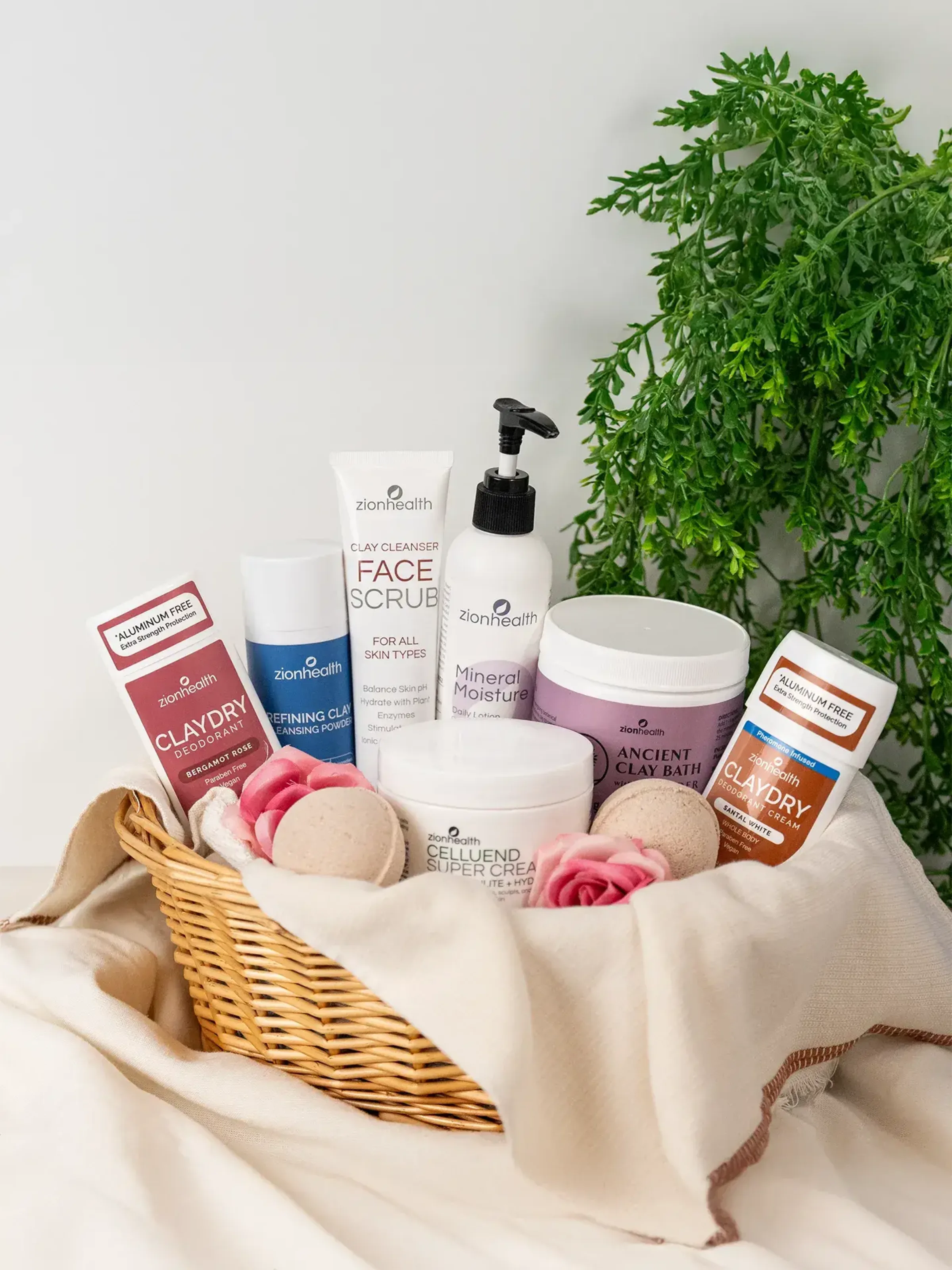 A wicker basket filled with Zion Health Claydry Deodorant, Refining Clay Cleansing Powder, Face Scrub, Mineral Moisture, Ancient Clay Bath, and Celluend Super Cream, surrounded by roses and greenery.