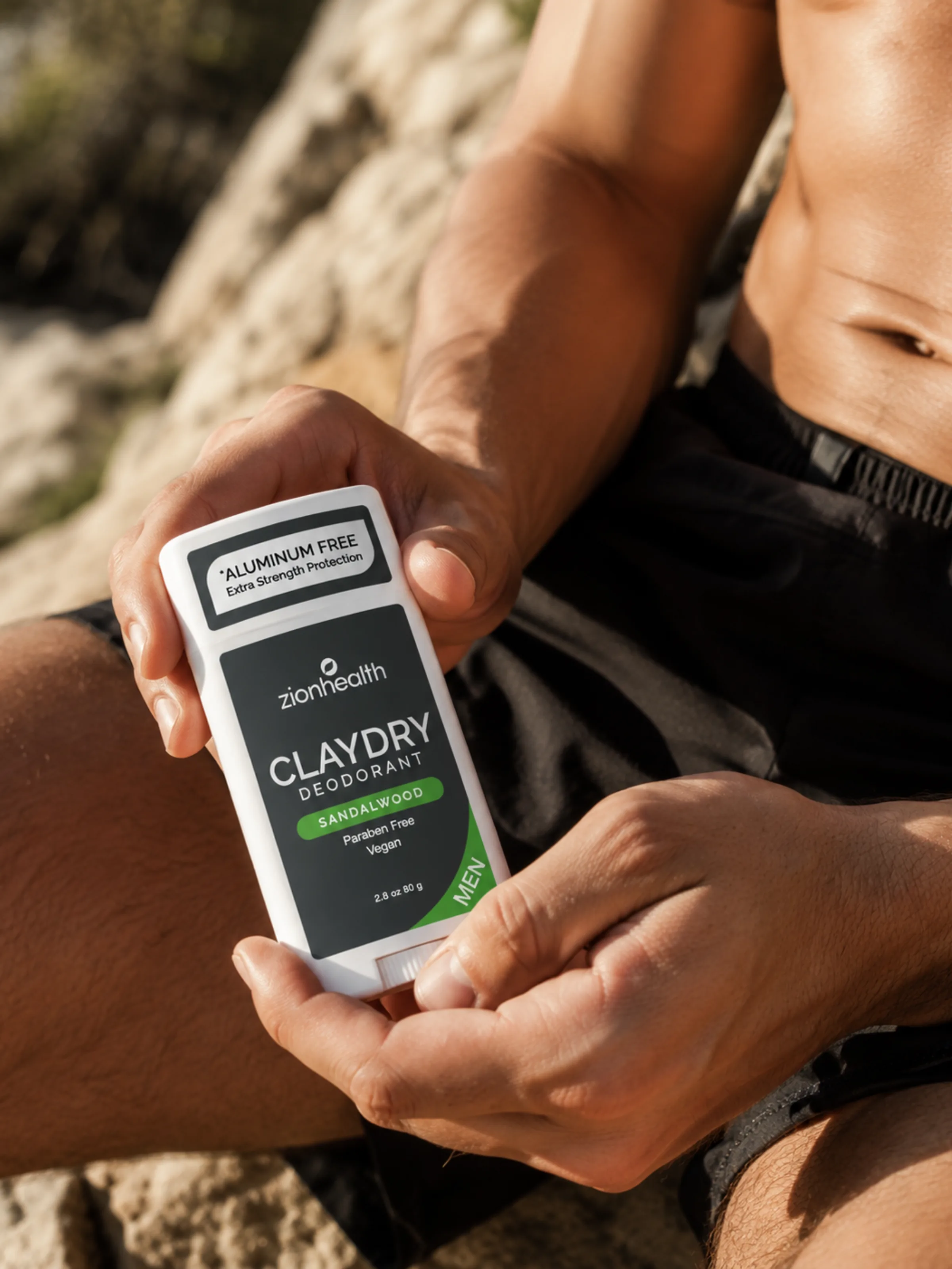 A man outdoors on rocks holding a Zion Health Claydry Deodorant Sandalwood Men stick.