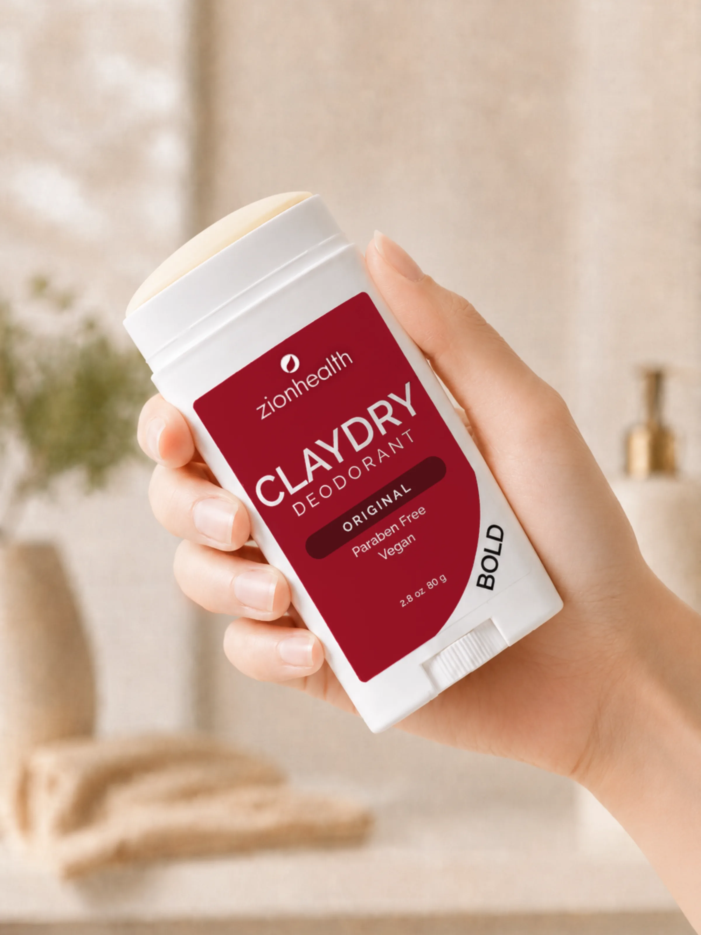 A hand holding Zion Health Claydry Deodorant Original Bold in a plant-filled bathroom.