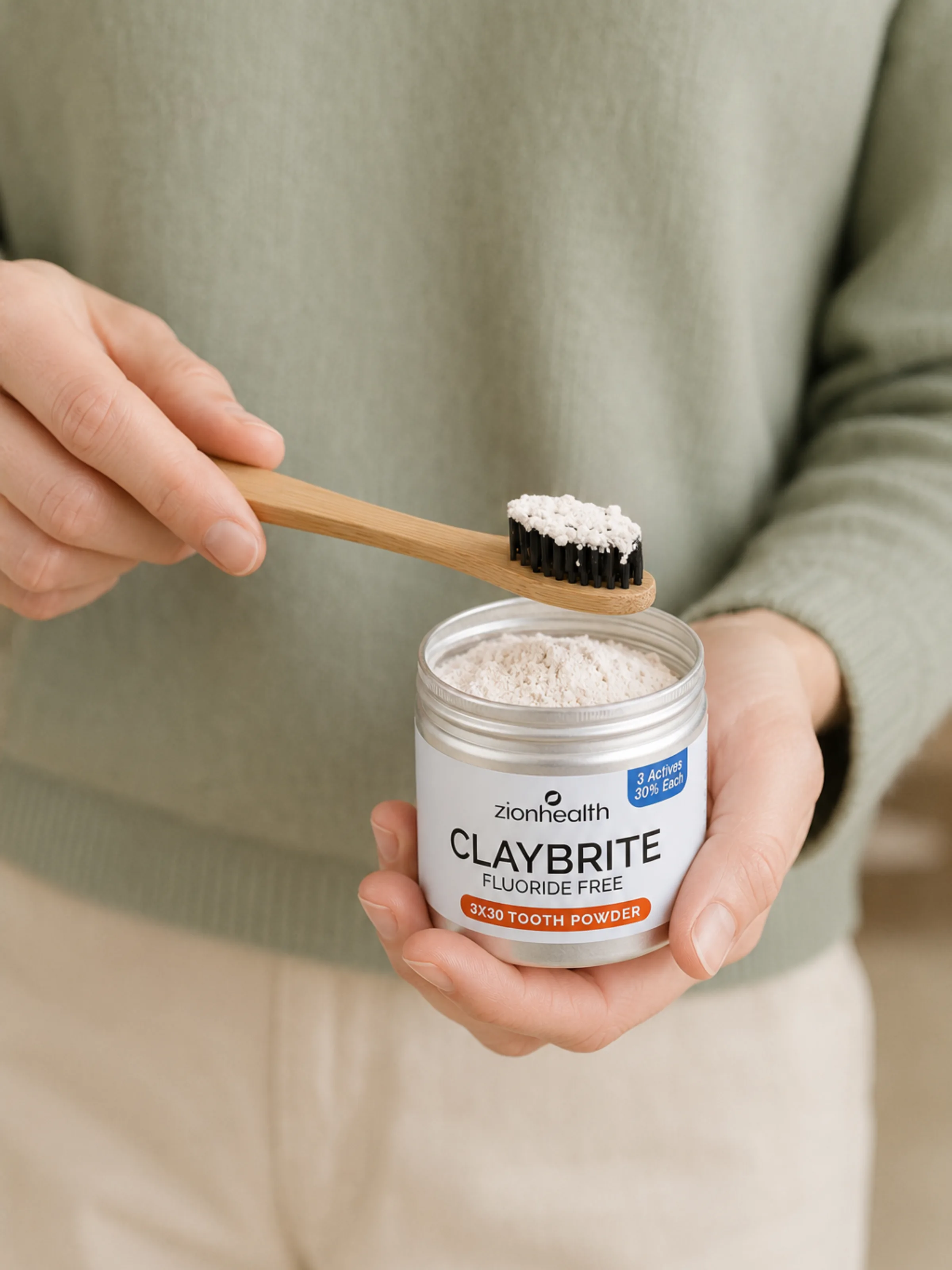 A bamboo toothbrush dipping into an open tin of Zion Health Claybrite tooth powder.