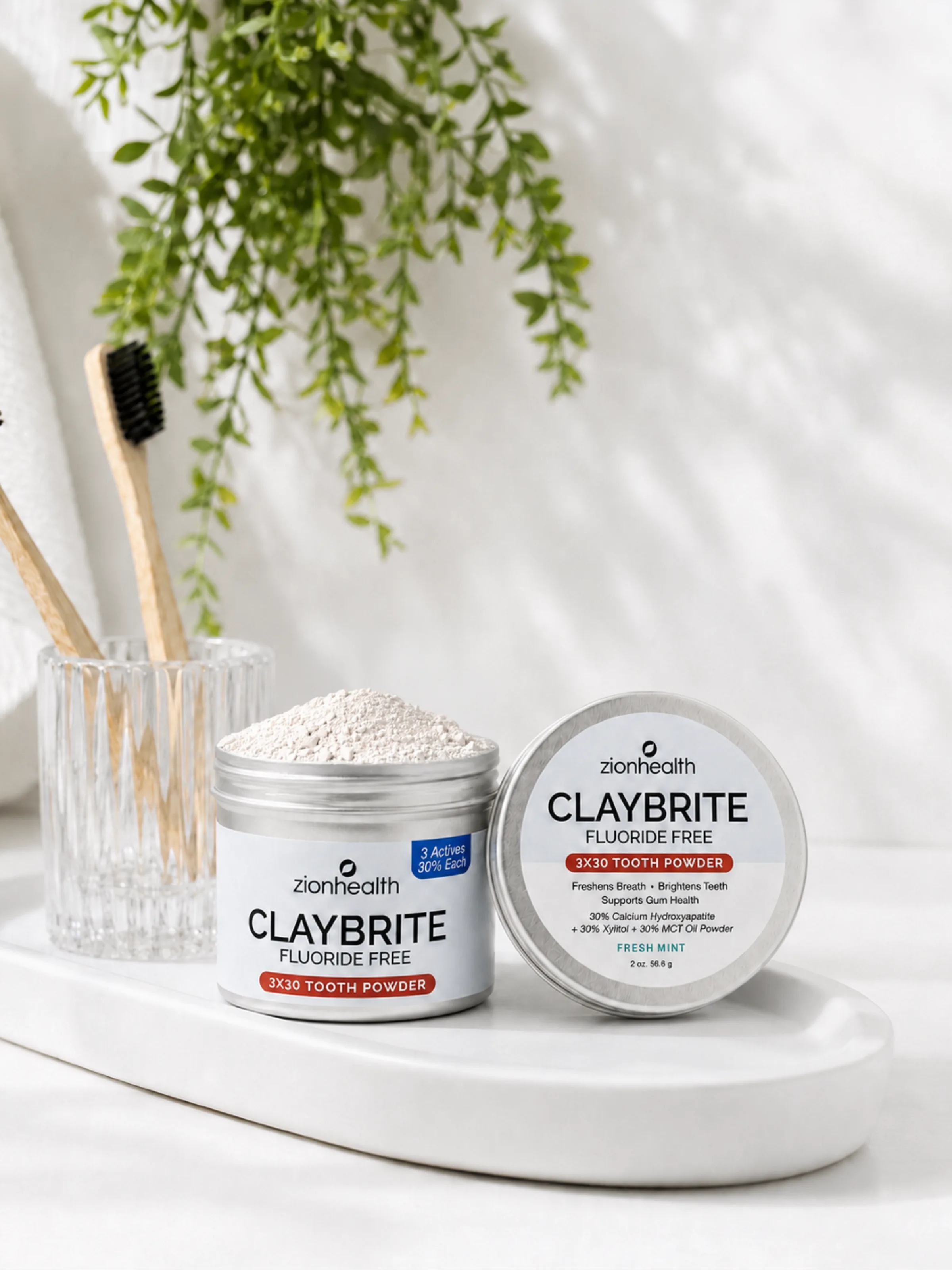 Two tins of Zion Health Claybrite Fluoride-Free tooth powder, one open and one closed, on a bathroom tray with toothbrushes.