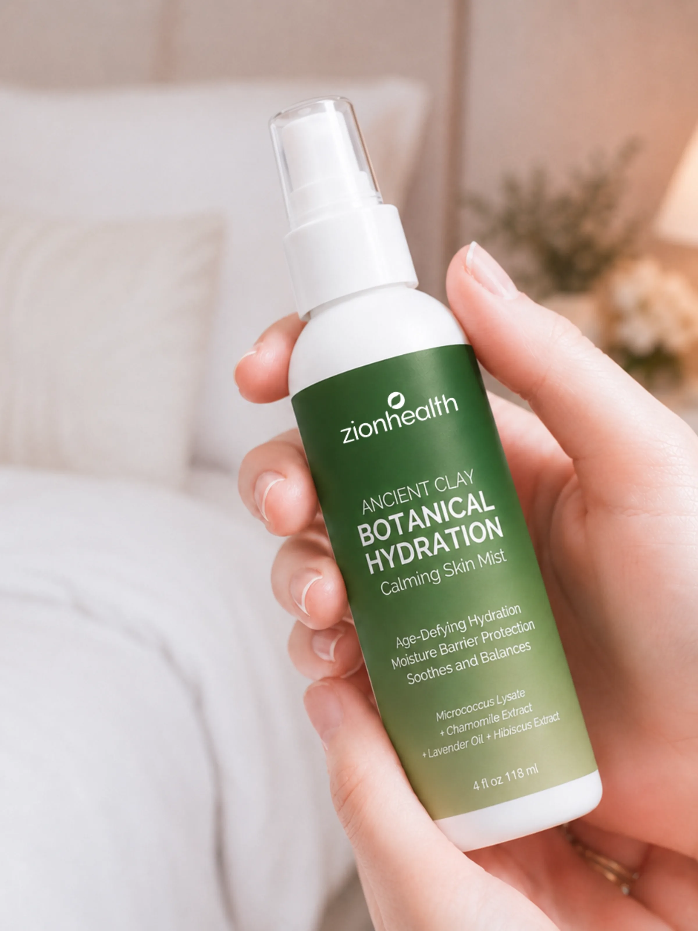 A hand holding Zion Health Ancient Clay Botanical Hydration Calming Skin Mist in a soft bedroom setting.