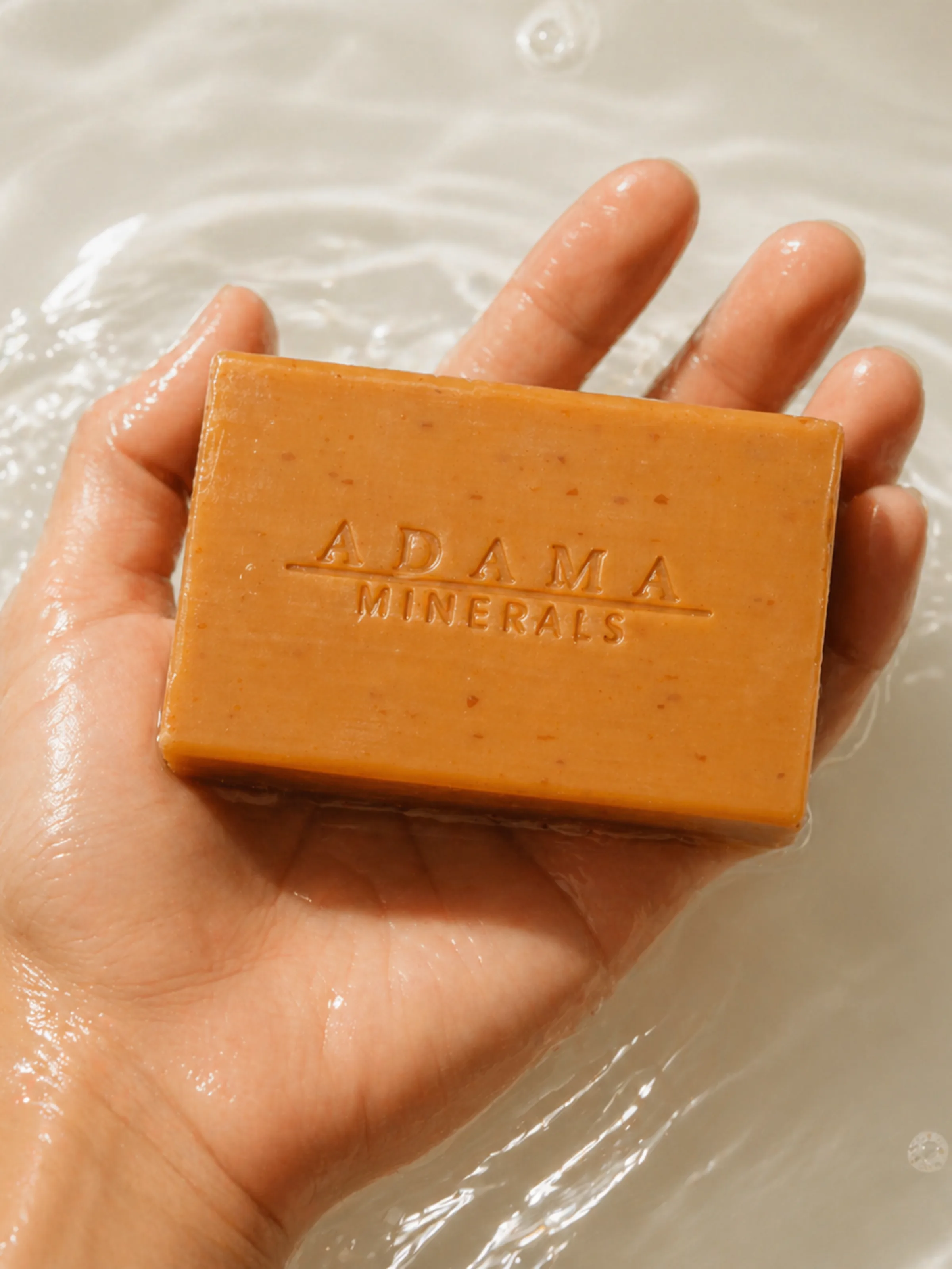 A hand holding an Adama Minerals clay soap bar in bathwater.
