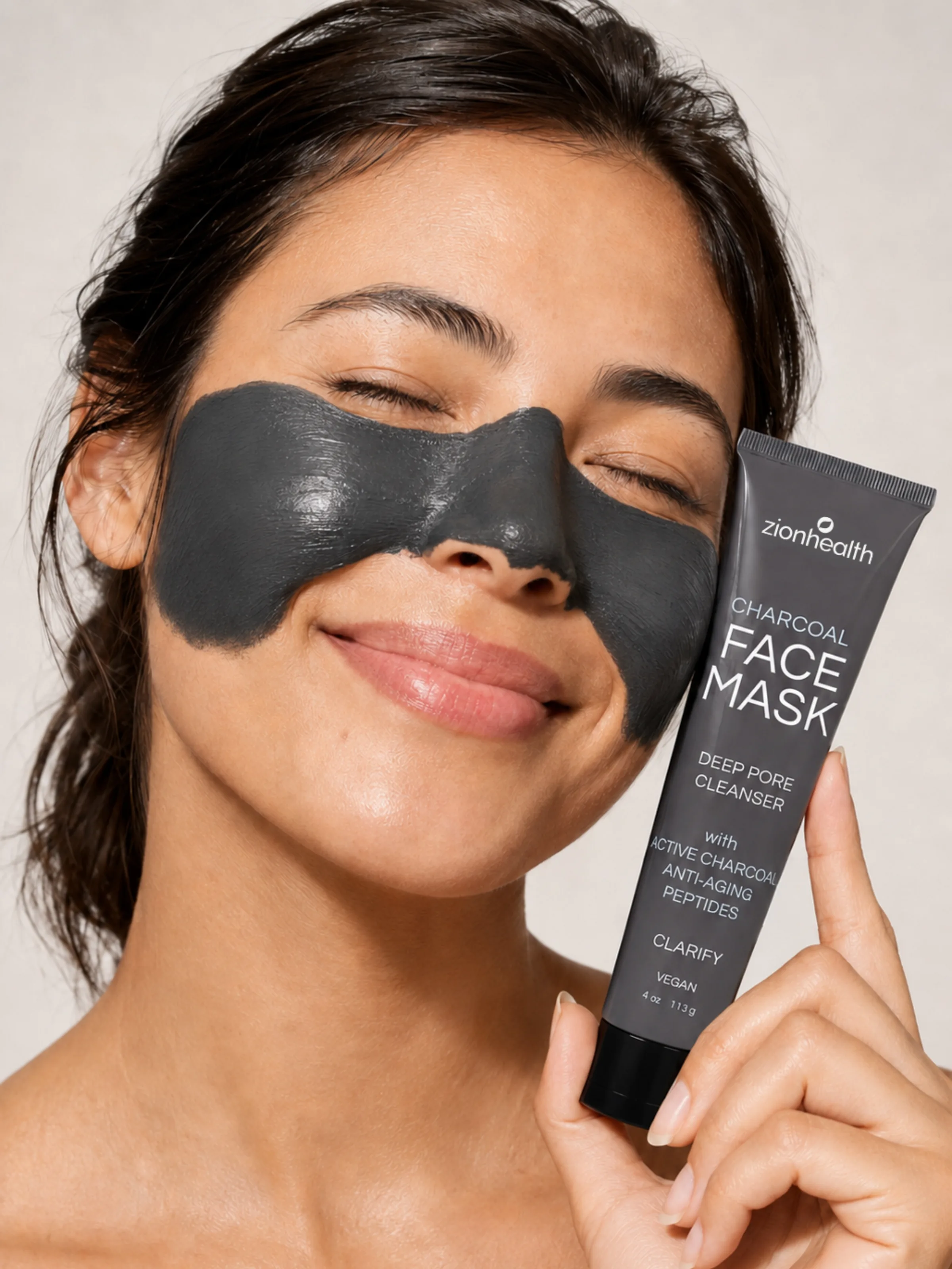 A woman with a charcoal mask on her face holding a tube of Zion Health Charcoal Face Mask.