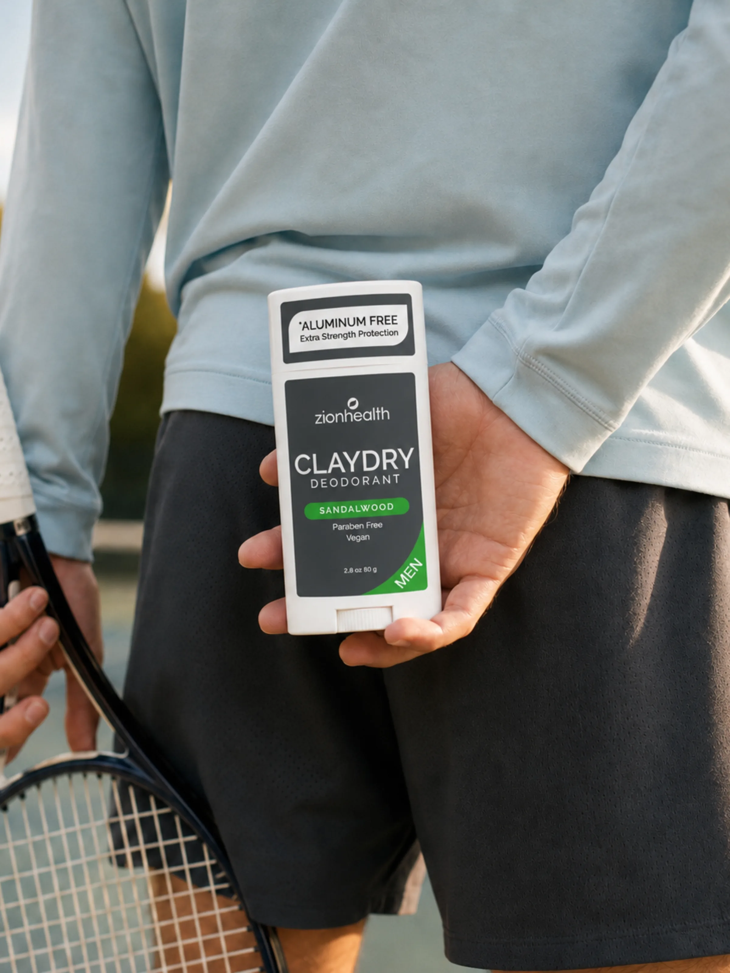 A man holding a Zion Health Claydry Deodorant Sandalwood Men stick behind his back on a tennis court.