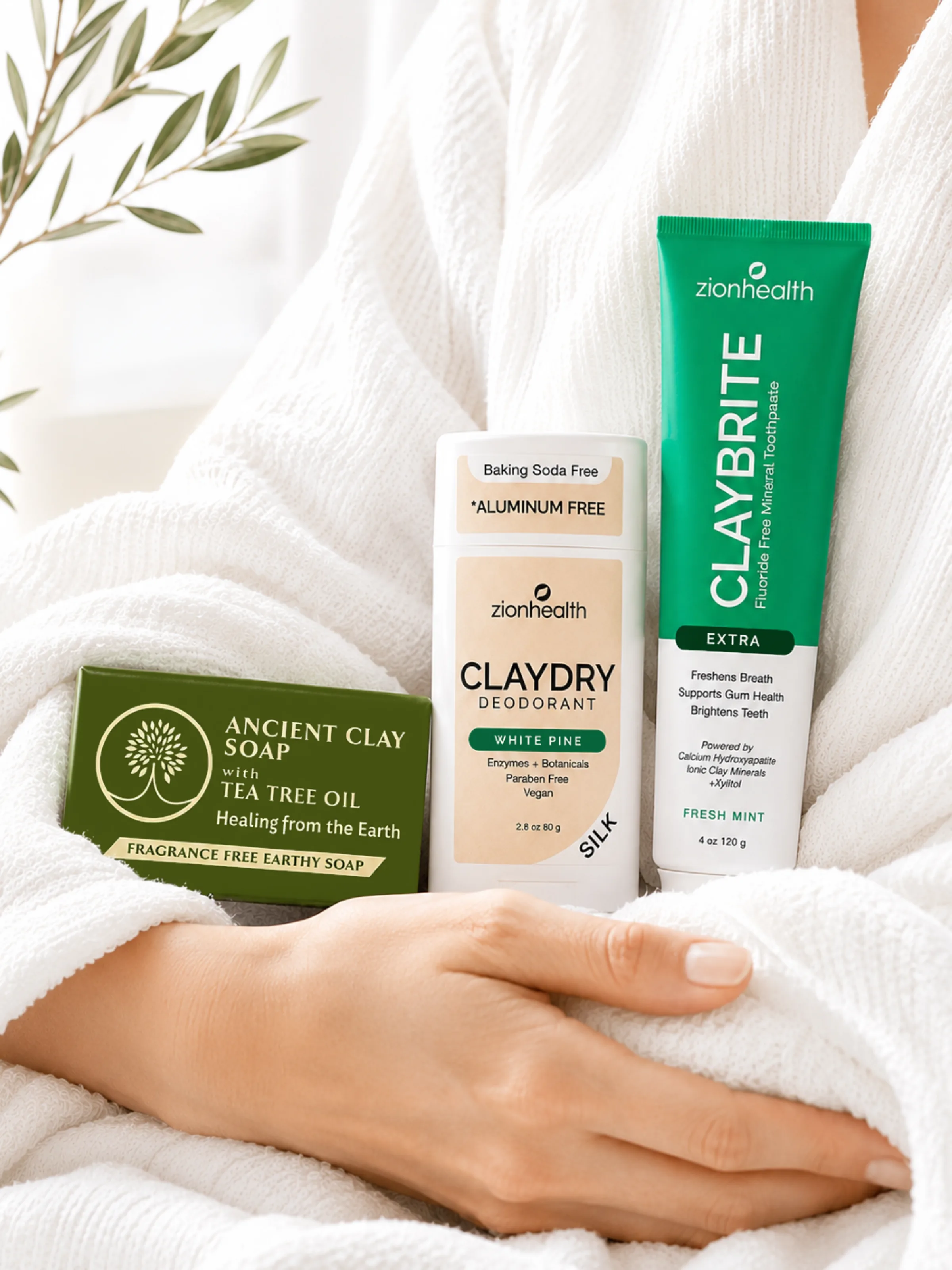 A basket of Zion Health products including Claydry Deodorant, Refining Clay Cleansing Powder, Face Scrub, Mineral Moisture, Ancient Clay Bath, and Celluend Super Cream, wrapped in linen with roses and bath bombs.
